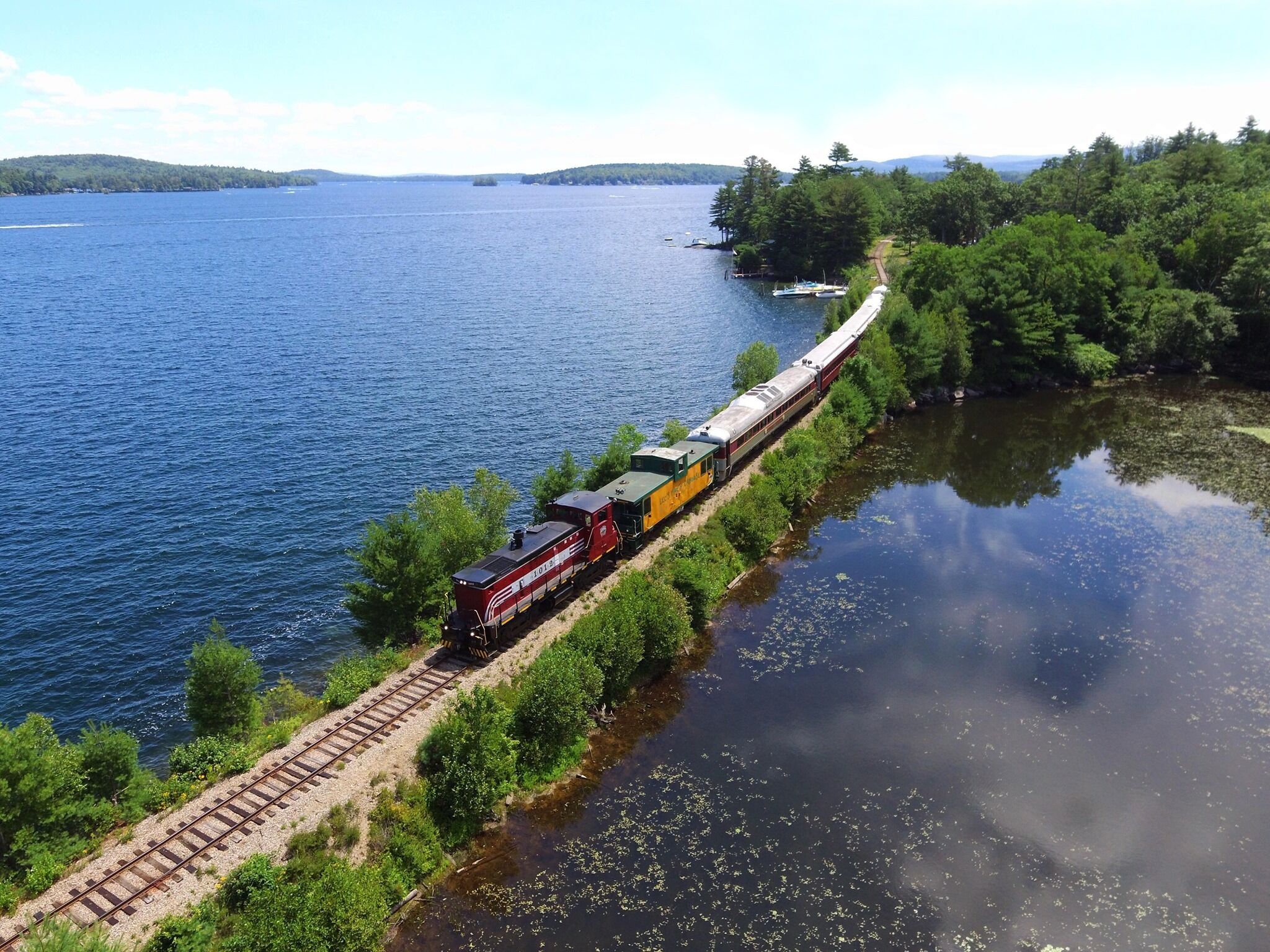 A train traveling along a narrow track beside a body of water, surrounded by dense greenery.