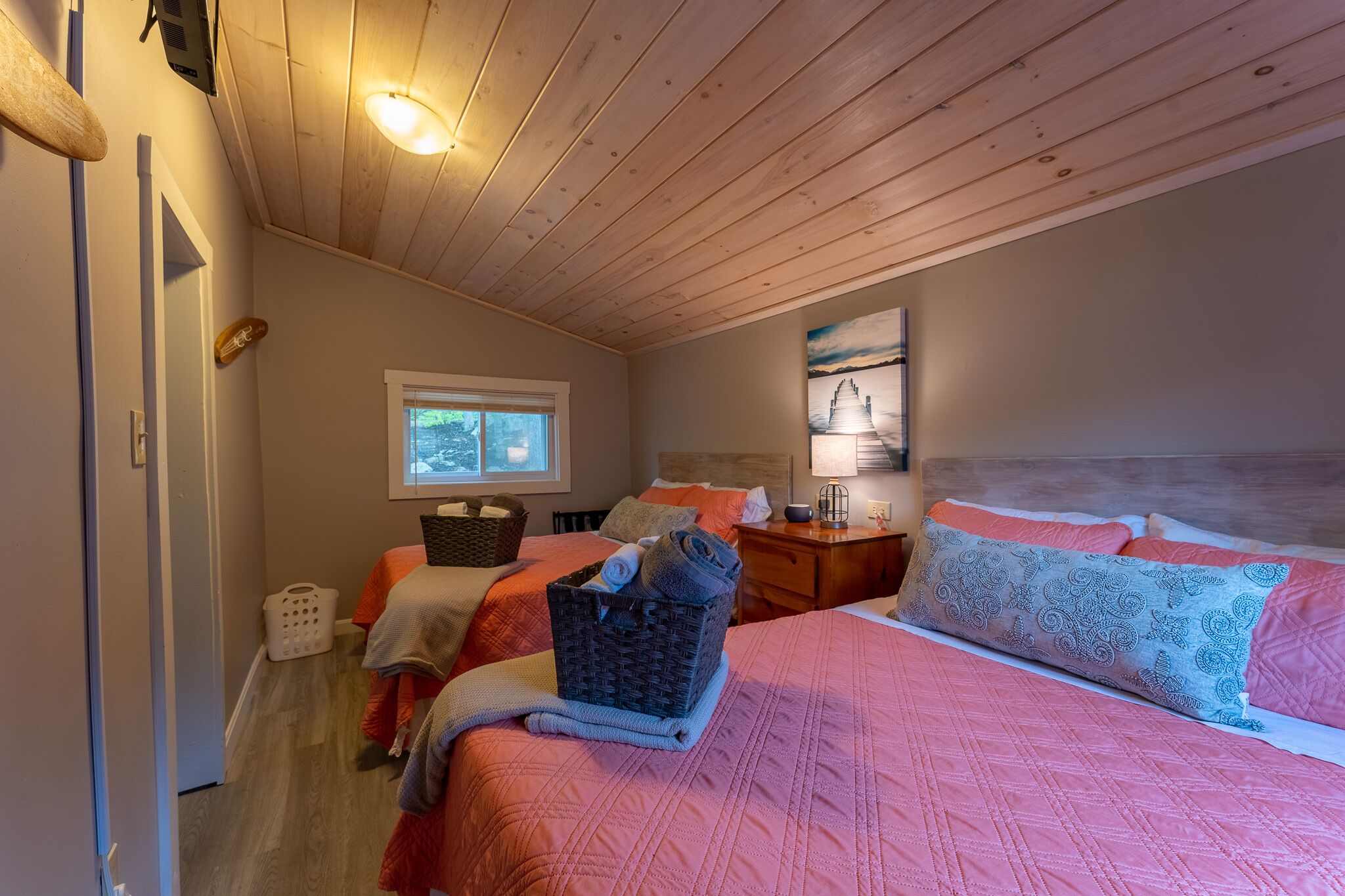 Ein gemütliches Zimmer mit Holzboden, einem Bett mit rosa Bettwäsche, einem Nachttisch mit Lampe und einem Fenster mit Blick auf Bäume.