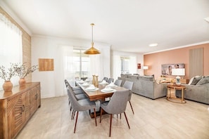 A modern living room with a dining area, grey sofas, a wooden dining table, and a pendant light.