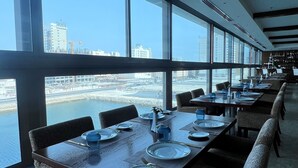 A restaurant with a long table set for dining, large windows offering a view of a river and buildings, and patterned chairs. - The Seven Hotel (Manama)