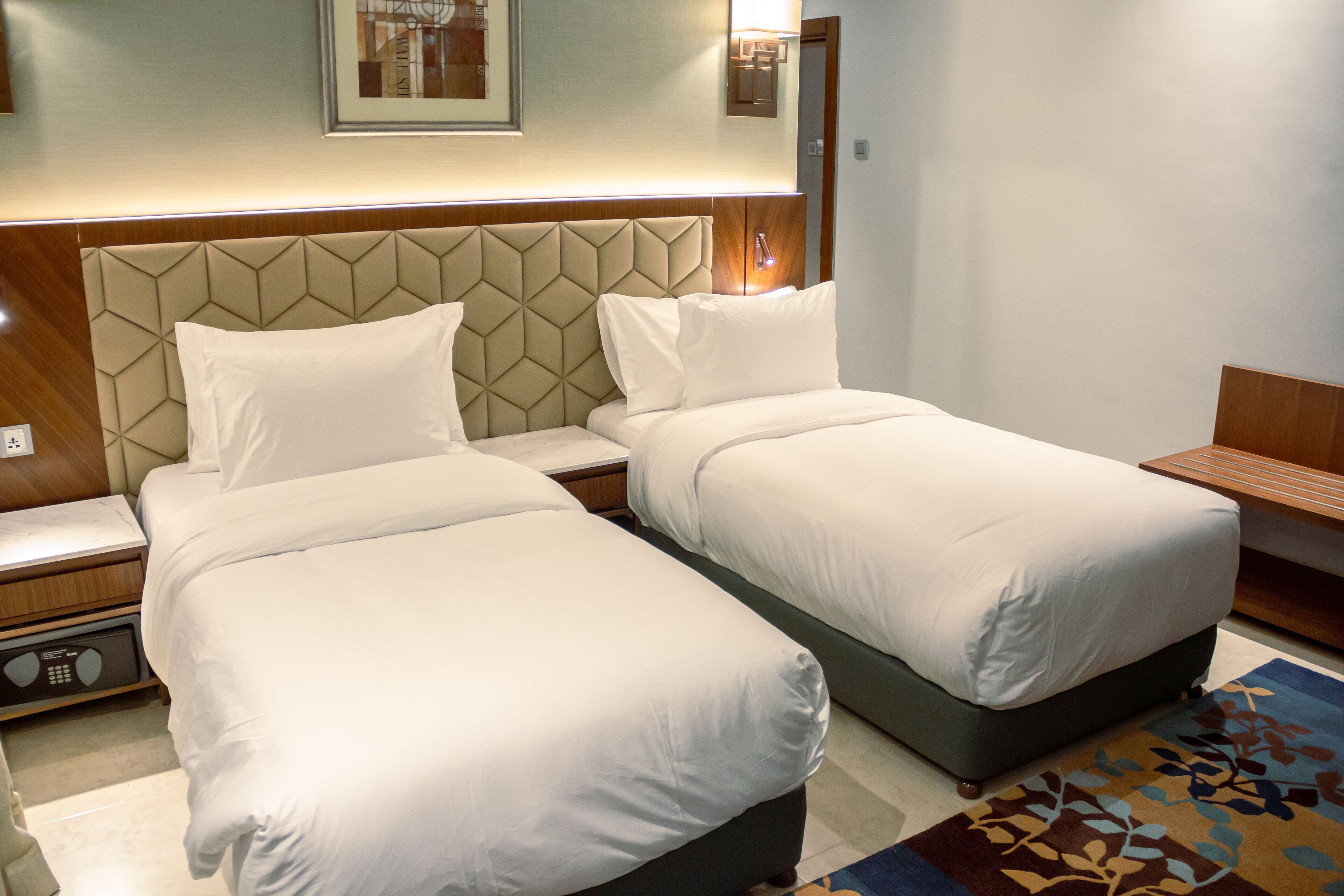 A hotel room with two beds, a wooden headboard, a bedside table, a nightstand, and a patterned rug.