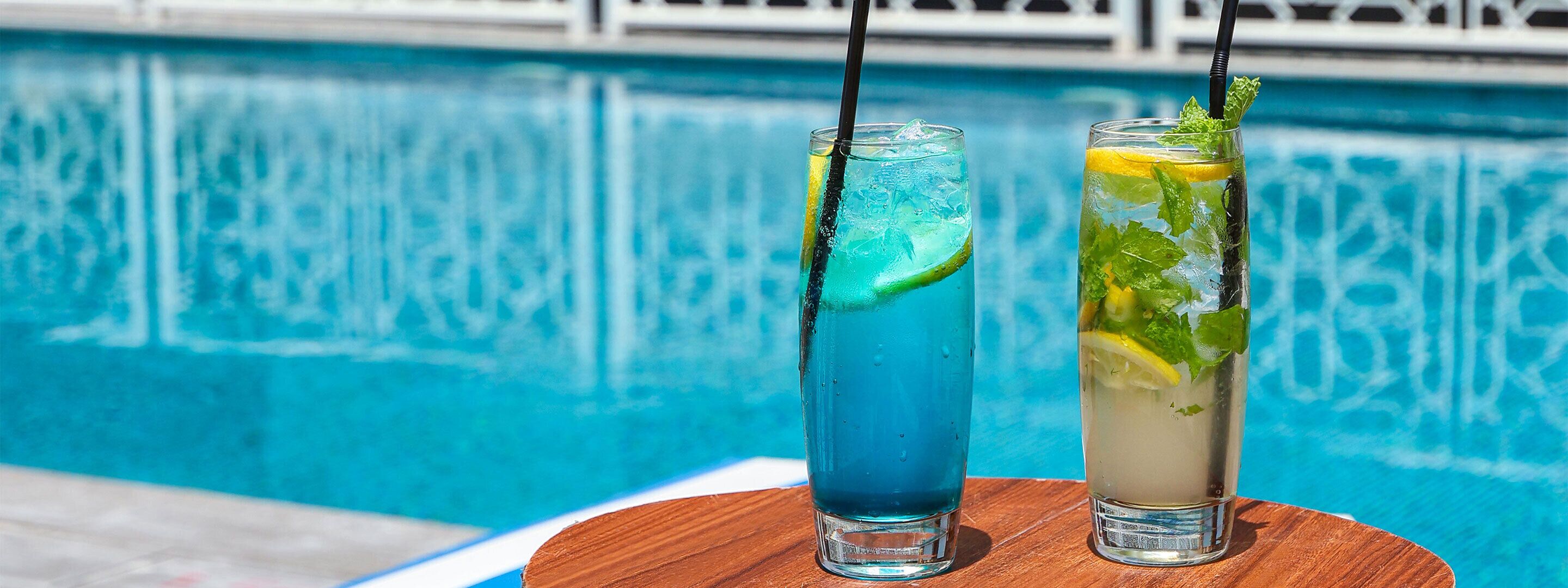 Dos vasos de bebidas con pajitas y adornos, sobre una superficie de madera junto a una piscina.