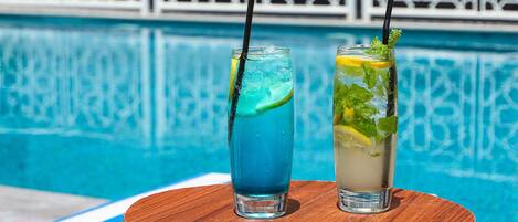 Two glasses of beverages with straws and garnishes, placed on a wooden surface by a pool.