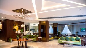 A modern hotel lobby with a wooden reception desk, a round table with vases, a green sofa, and a large TV screen. - The Seven Hotel (Manama)