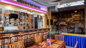 Zona de bar con sillas y mesas de madera, una barra con estantes llenos de botellas y un rústico muro de piedra de fondo.