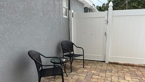 En uteplats med två stolar och ett bord, ett vitt staket och en grå vägg.