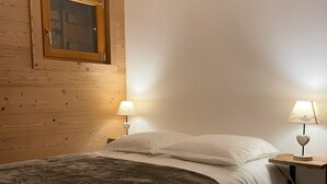 A bedroom with a wooden headboard, a window with a wooden frame, two bedside lamps, a bed with white and brown bedding, and a wooden floor.