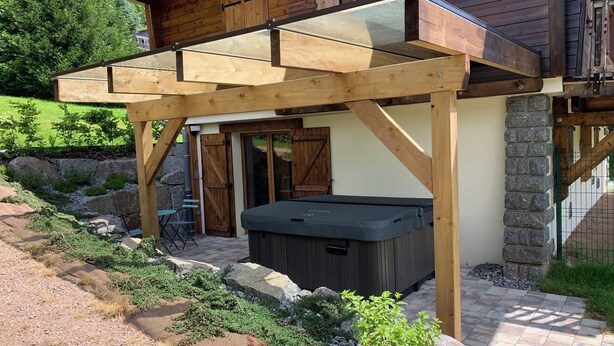 A wooden cabin with a glass-enclosed patio, a hot tub, and a stone wall.