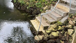 Un puente de piedra sobre un río tranquilo, rodeado de vegetación y una escalera de metal que desciende hacia el agua.