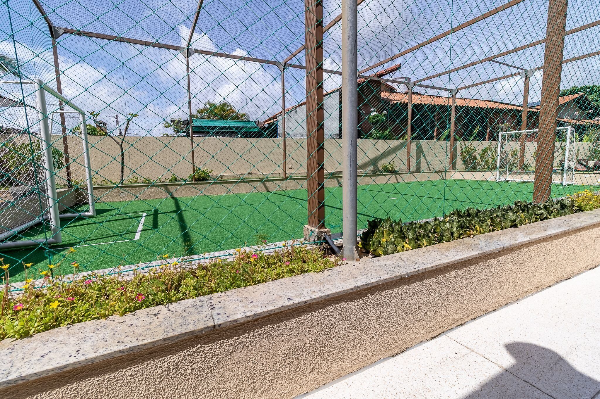 A fenced sports court with a green surface, surrounded by a low hedge and a concrete pathway.