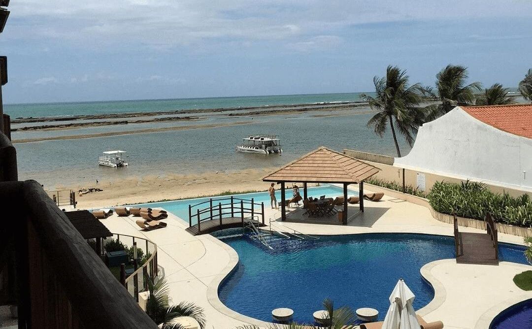 A pool area with a view of the beach and ocean, a boat on the water, and a covered seating area.