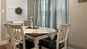 Un comedor con una mesa dispuesta para cuatro personas, rodeada de sillas blancas con cojines oscuros. Un mantel blanco cubre la mesa, y un pequeño jarrón con flores está colocado sobre ella.
