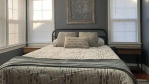 Un dormitorio con cama, dos mesitas de noche, un adorno en la pared y ventanas amplias con persianas.