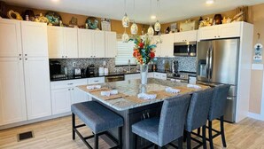 Una cocina moderna con armarios blancos, una isla central con taburetes de bar, electrodomésticos de acero inoxidable y objetos decorativos en los gabinetes superiores.