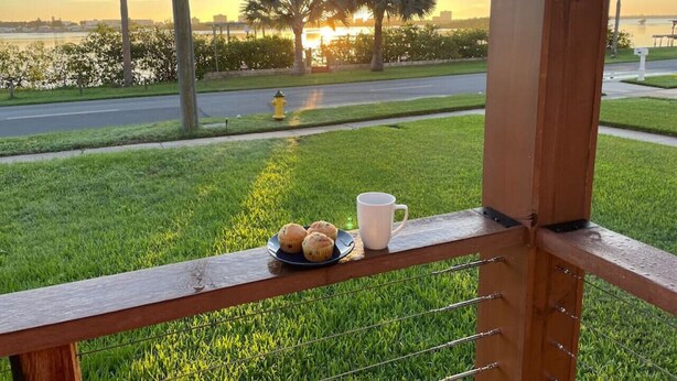 Vista desde un balcón con un plato de muffins y una taza, con vistas a un área verde con palmeras y un atardecer sobre el agua.