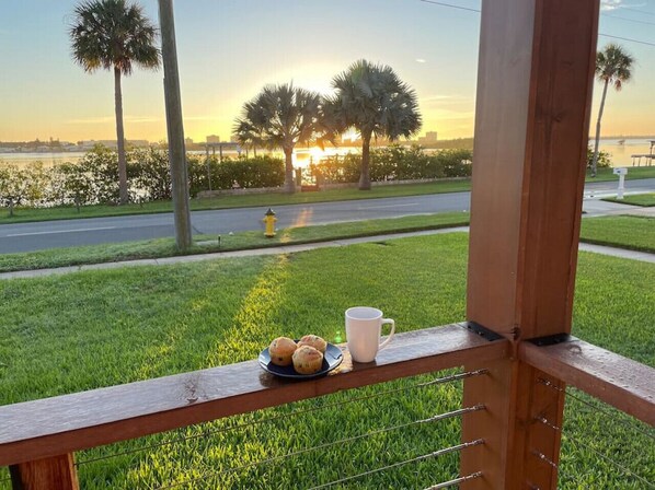 A view from a balcony with a plate of muffins and a mug, overlooking a grassy area with palm trees and a sunset over a body of water.