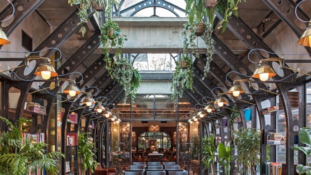Un restaurant avec un plafond haut, des plantes suspendues et un espace central aménagé avec des tables et des chaises.