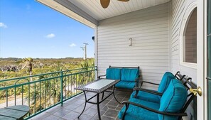 A covered balcony with a table and two chairs, overlooking a landscape with vegetation and a clear sky.