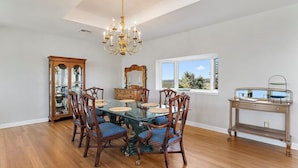 A dining room with a glass-top table, wooden chairs, a chandelier, a wooden cabinet, and a window with a view. - Carpe Beachiem Home, Ocean Views & Dual Decks (Ponte Vedra Beach)