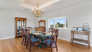 A dining room with a glass-top table, wooden chairs, a chandelier, a wooden cabinet, and a window with a view. - Carpe Beachiem Home, Ocean Views & Dual Decks (Ponte Vedra Beach)