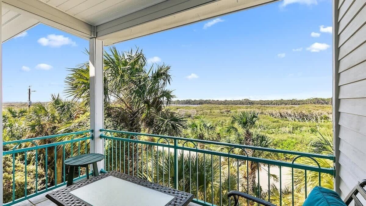 A covered balcony with a table and chairs, overlooking a natural landscape with palm trees and a distant horizon.