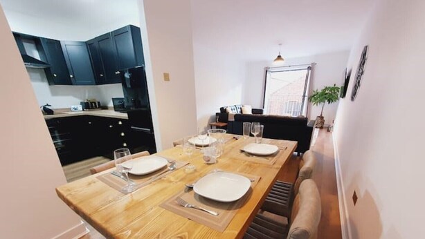 A dining area with a wooden table set for four, a kitchen in the background, and a window with a view. - Best Location in Belfast! Free Parking-Luxury Accommodation (Belfast)