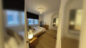 Un dormitorio con cama, mesita de noche, lámpara, cómoda y vista de la habitación contigua a través de un espejo.