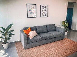 A grey sofa with two pillows, a framed artwork above it, and a potted plant to the left.