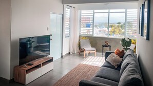 En moderne stue med et fladskærms-tv på et træstel, en grå sofa, en loftventilator og et stort vindue med udsigt over byen.