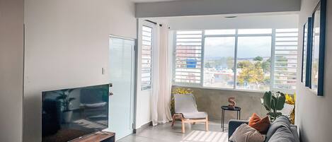 Una sala de estar moderna con un televisor de pantalla plana sobre un soporte de madera, un sofá gris, un ventilador de techo y un ventanal con vistas a la ciudad.