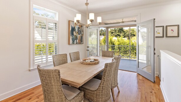 A dining area with a wooden table and wicker chairs, a chandelier, and a view of greenery through large windows. - Beach Chickens is your vacation destination for Pool Side Relaxation (St. Simons Island)
