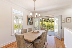 Un coin repas avec une table en bois et des chaises en osier, un lustre et une vue sur la verdure grâce à de grandes fenêtres.