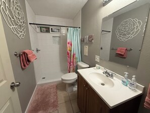 Un baño con inodoro, lavamanos y ducha. Hay toallas en la pared y una alfombra rosa en el suelo.