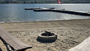 Una playa con un muelle de madera y una bandera roja, un pozo para fuego y montañas al fondo.