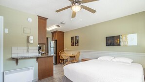 Habitación de hotel con cama, escritorio y silla, ventilador de techo y zona de kitchenette.