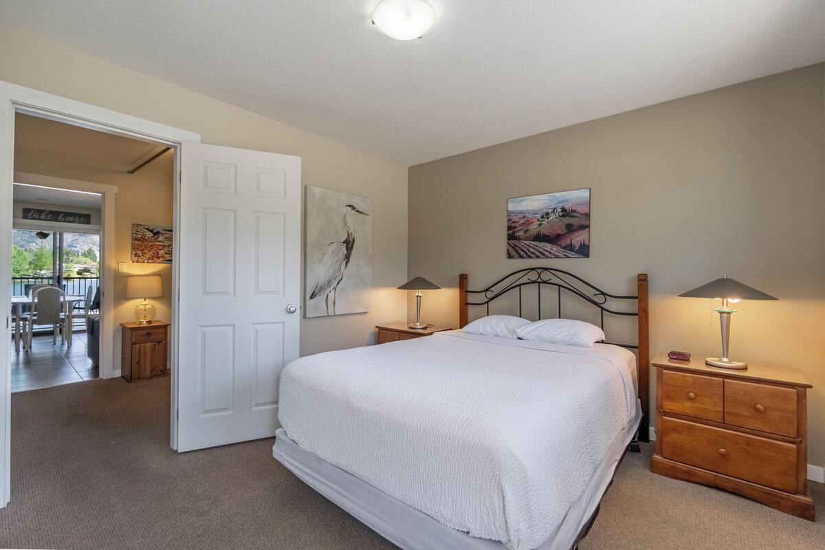 Un dormitorio con una cama grande, dos mesitas de noche con lámparas, un aparador de madera y una puerta que da a un comedor.