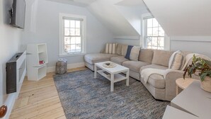 Un appartement moderne de style « loft », avec un plafond haut, une grande fenêtre, un canapé beige, une table basse blanche et un téléviseur à écran plat fixé au mur.