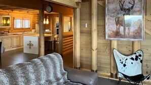 En rustik stuga med en inredning som inkluderar en matplats med träbord, en mysig sittgrupp med en mjuk fäll och en stor tavla med ett rådjur på väggen.