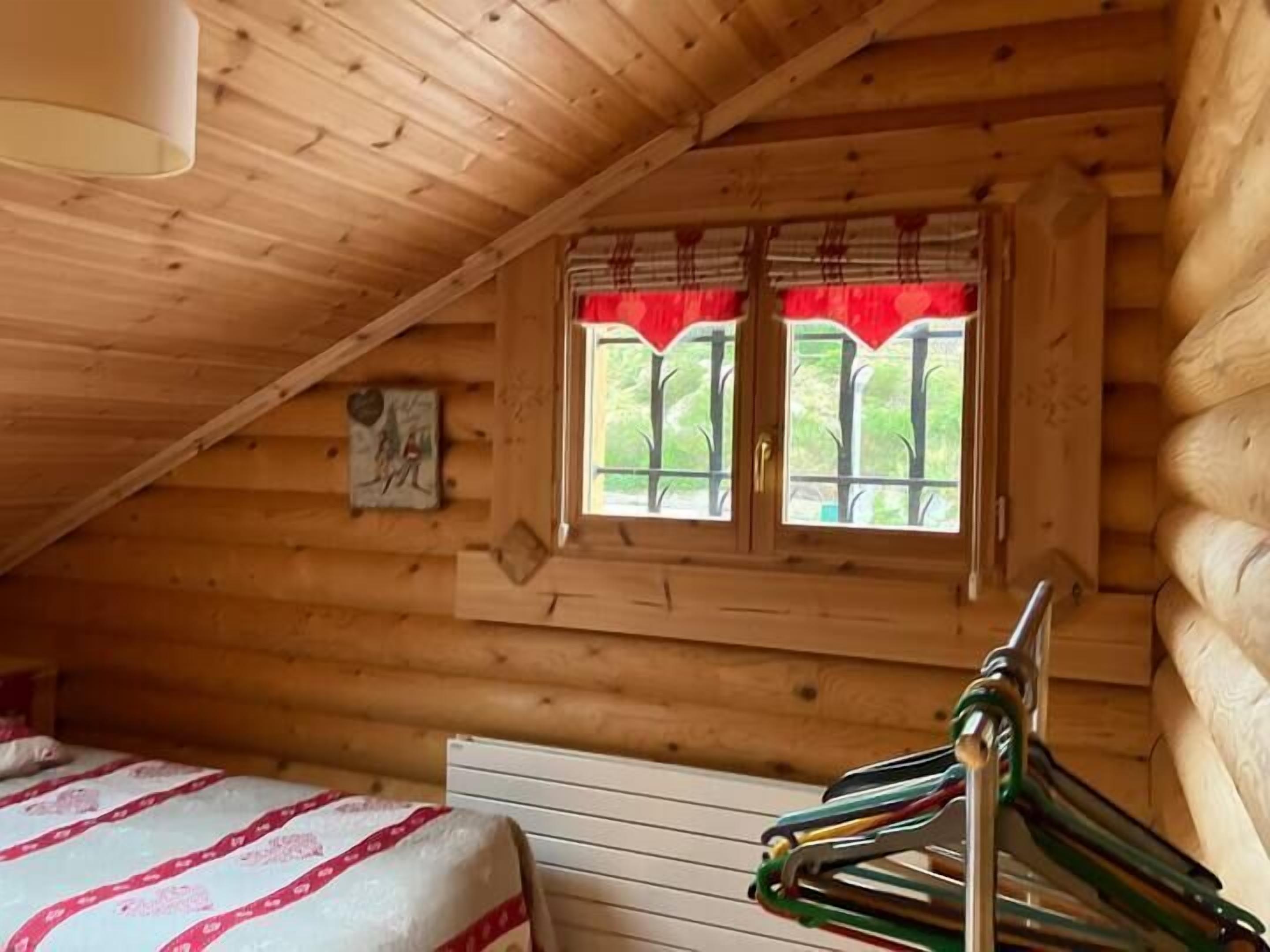 A wooden cabin room with a bed, a window with red curtains, and a wooden bench.