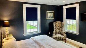 A bedroom with a bed, two windows with blinds, a chair, a lamp, and a framed picture on the wall. - Coastal Home in Prime location! Sleeps 14! (Marblehead)
