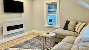 Un salon moderne avec un canapé beige, un téléviseur à écran plat fixé au-dessus d’une cheminée et une petite table ronde.
