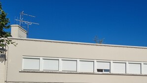 Un edificio con fachada plana, varias ventanas con contraventanas y un coche estacionado adelante.