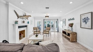 A spacious living room with a fireplace, a sofa, a coffee table, and a dining area. There is a view through the glass doors to the outside.