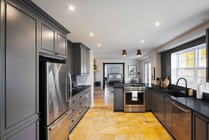 Kitchen features sleek cabinetry, a stainless steel refrigerator, and a central island with a sink, leading to a bedroom with a bed and outdoor views.