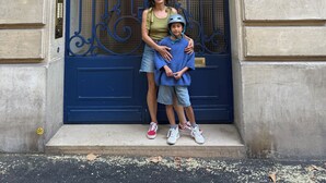 Una mujer y un niño frente a una puerta azul con detalles de metal ornamentales, una placa con el número '6' y una fachada de edificio de piedra.