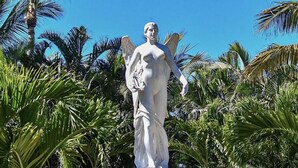 Una estatua de una figura alada, rodeada de palmeras y un cielo azul despejado.