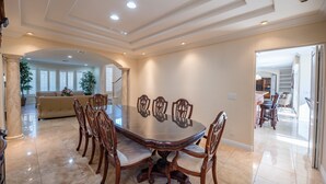 A spacious dining room with a large oval table and six chairs, a kitchen area visible through an open doorway, and a living room with a sofa and potted plants. - Large Luxurious Estate on Golf Course w/pool/spa in Gated Community (Henderson)