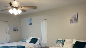 Ein Schlafzimmer mit zwei Betten, jeweils mit weißer Bettwäsche und türkisfarbenen Kissen, einem Deckenventilator und Kunstwerken an den Wänden.