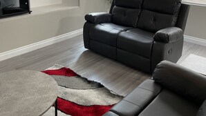 Ein modernes Wohnzimmer mit grauem Ledersofa, einem flachbildfernseher an der Wand und einem großen Fenster mit Jalousien.