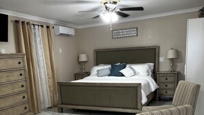 Un dormitorio con cama, sillón, cómoda, mesita de noche, lámpara, ventilador de techo y aire acondicionado instalado en la pared.